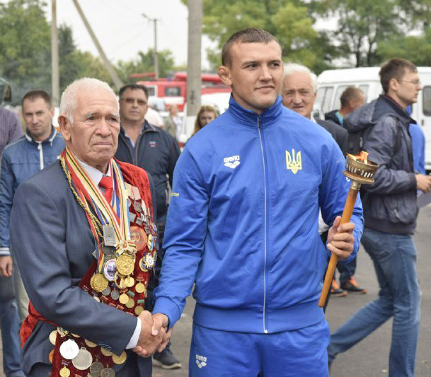 Запалив вогонь змагань чемпіон України серед юніорів борець Євген Совєта (праворуч), отримавши його з рук олімпійського чемпіона 1964 року Андрія Хіміча (ліворуч). Фото з сайту ck-oda.gov.ua